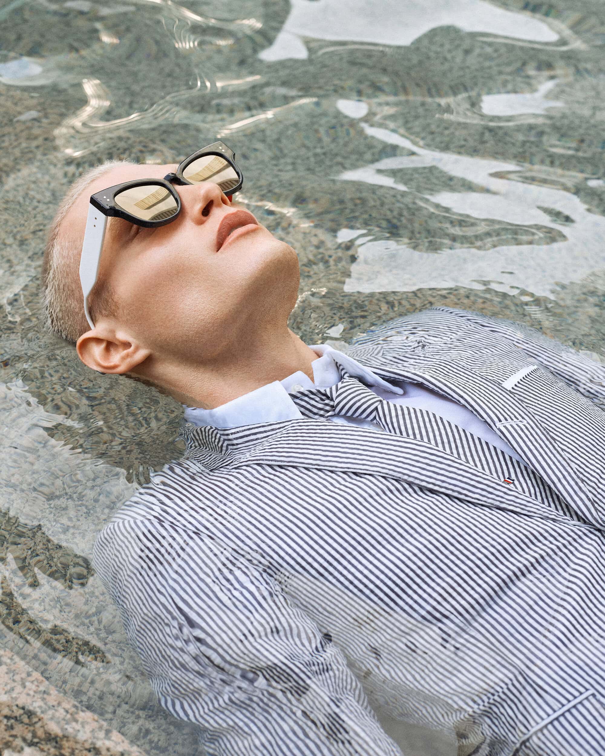 Male fashion model with blonde hair and sunglasses, wearing a blue and white striped suit, partially submerged in clear water outdoors.