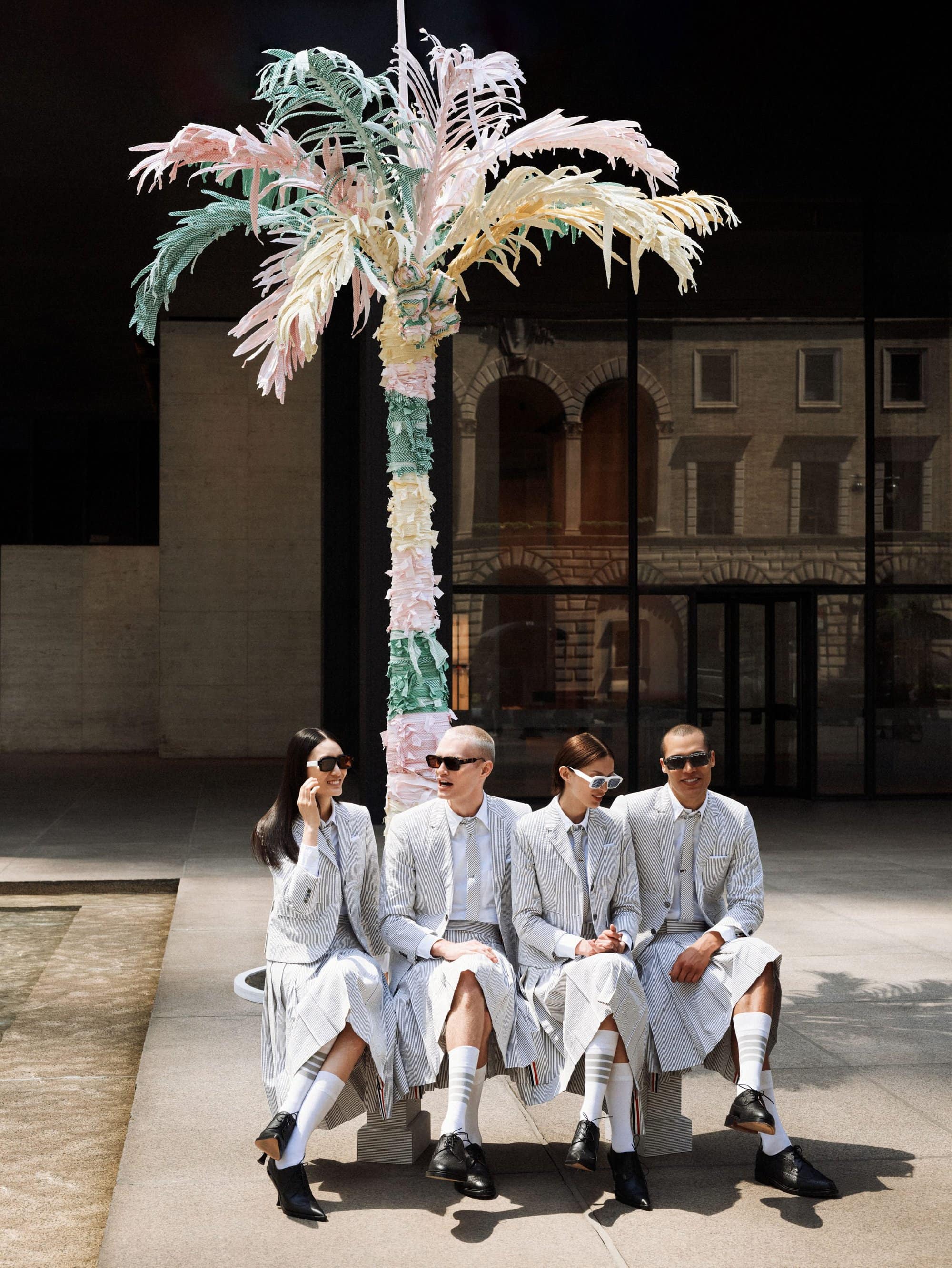 Four people in matching grey suits sit outdoors on a bench next to an artificial palm tree. Urban scene with modern architecture.