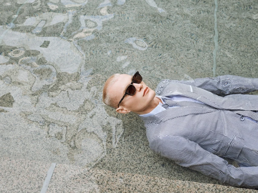 Adult male in sunglasses and pinstripe suit lies back in shallow water. Visible water reflections, sunlight, pale complexion.