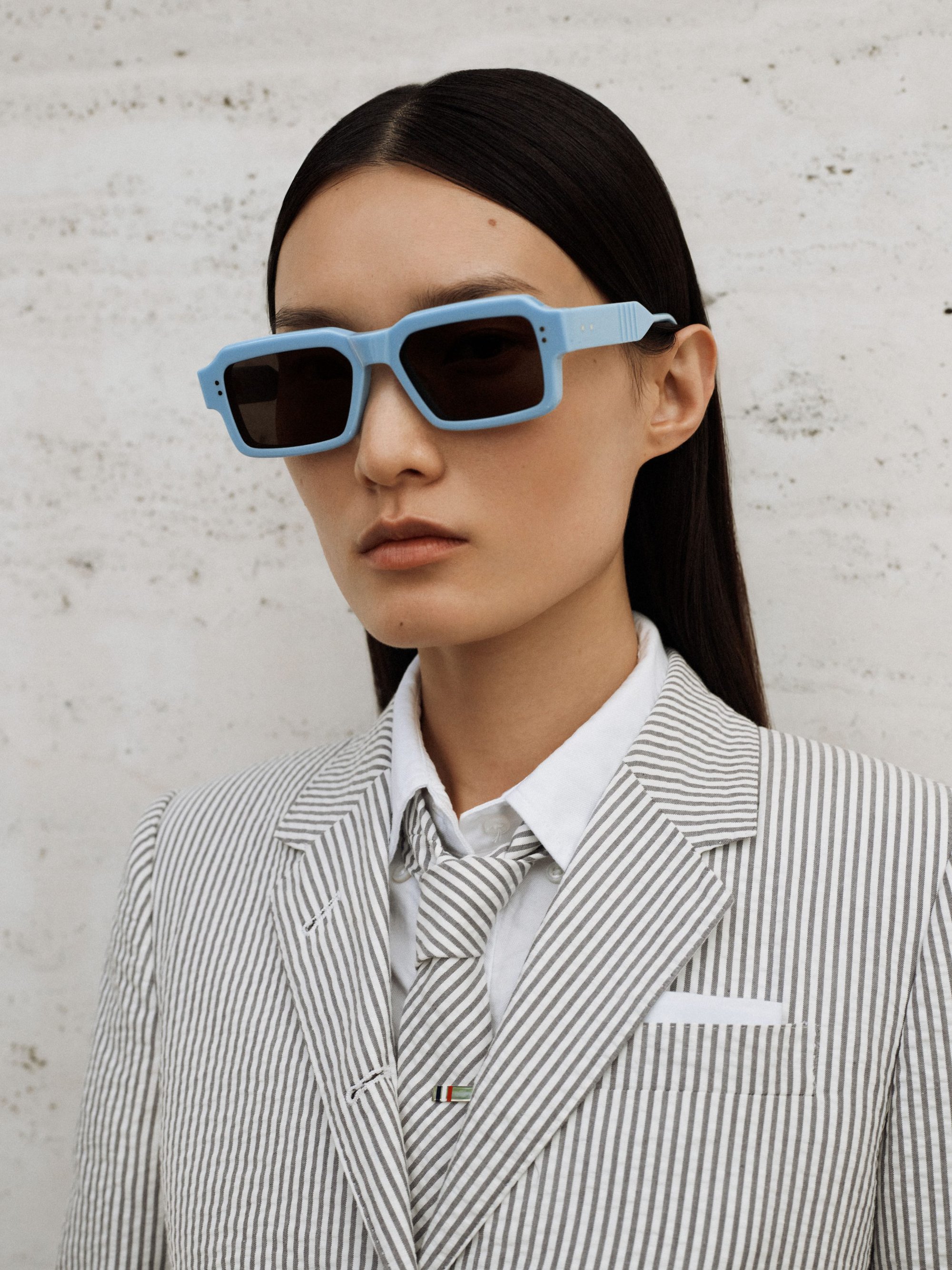 Portrait of a stylish woman wearing light blue sunglasses, a striped blazer, and tie against a textured, neutral background.