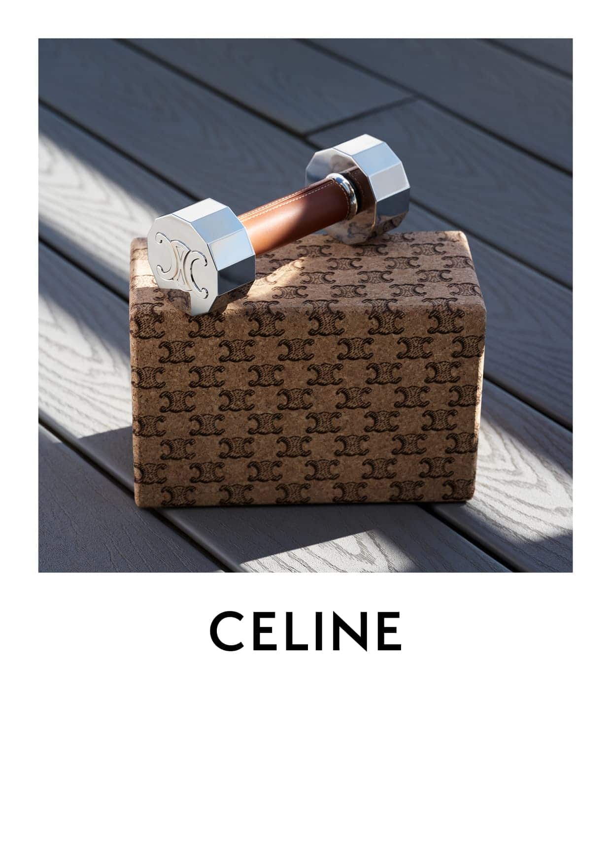 A dumbbell from Celine sits on a box patterned with the Celine logo, set against a neutral grey wooden background with natural light.