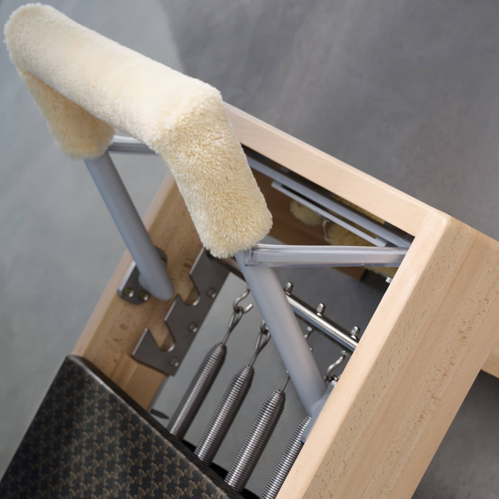 Pilates reformer with a light wood frame, metal springs, and a cream-colored furry pad on the headrest against a grey background.