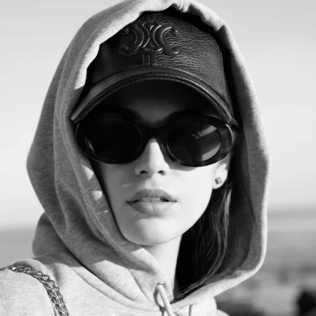 Close-up of a person in a grey hoodie, black leather baseball cap with branding, and dark oval sunglasses, shot in black and white.