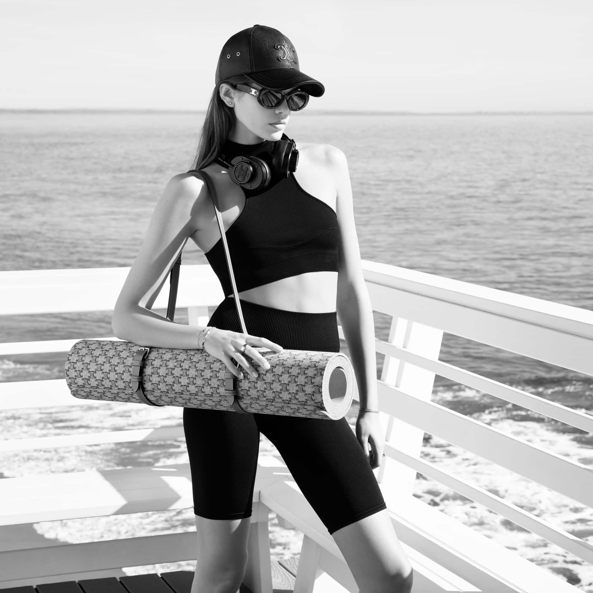 Athletic woman in workout clothes stands outdoors near the water holding a yoga mat, wearing sunglasses and a baseball cap.