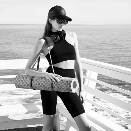 Athletic woman in workout clothes stands outdoors near the water holding a yoga mat, wearing sunglasses and a baseball cap.