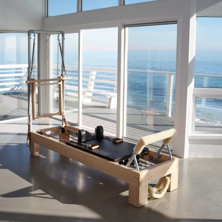 Pilates reformer machine on a concrete floor in a sunlit room with a view of the ocean and white wooden deck outside.