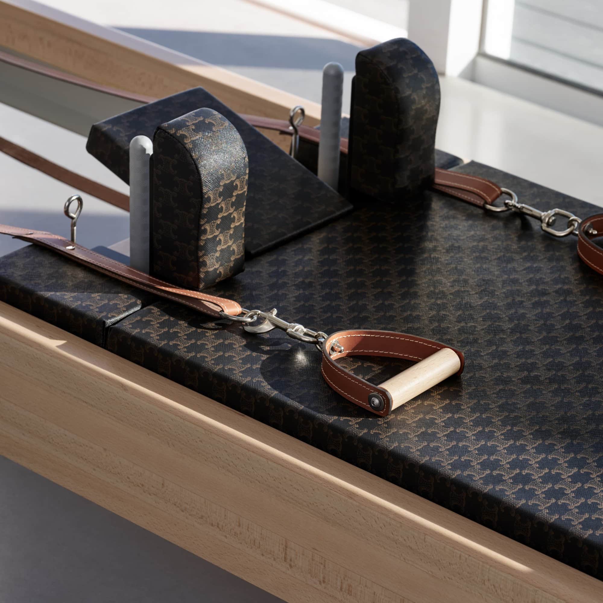 Black Pilates reformer with patterned fabric, leather straps, and silver hardware in a brightly lit indoor space. Fitness equipment.