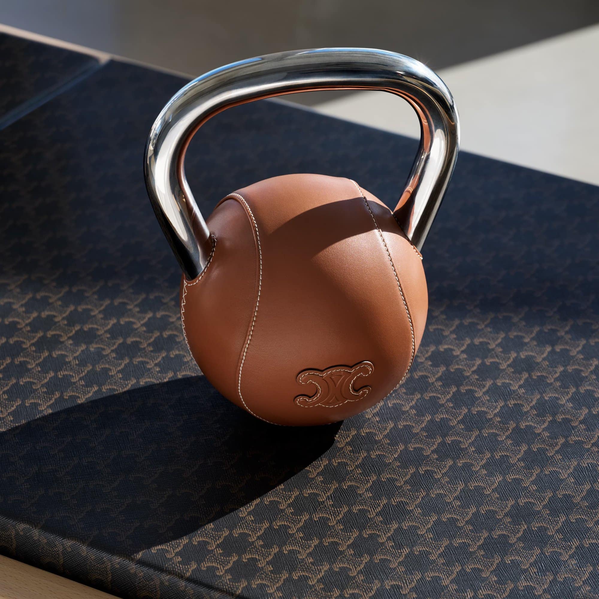 Close-up studio shot of a brown leather kettlebell with a shiny metal handle, adorned with the Celine logo, resting on a patterned surface.