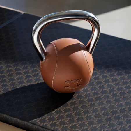 Close-up studio shot of a brown leather kettlebell with a shiny metal handle, adorned with the Celine logo, resting on a patterned surface.