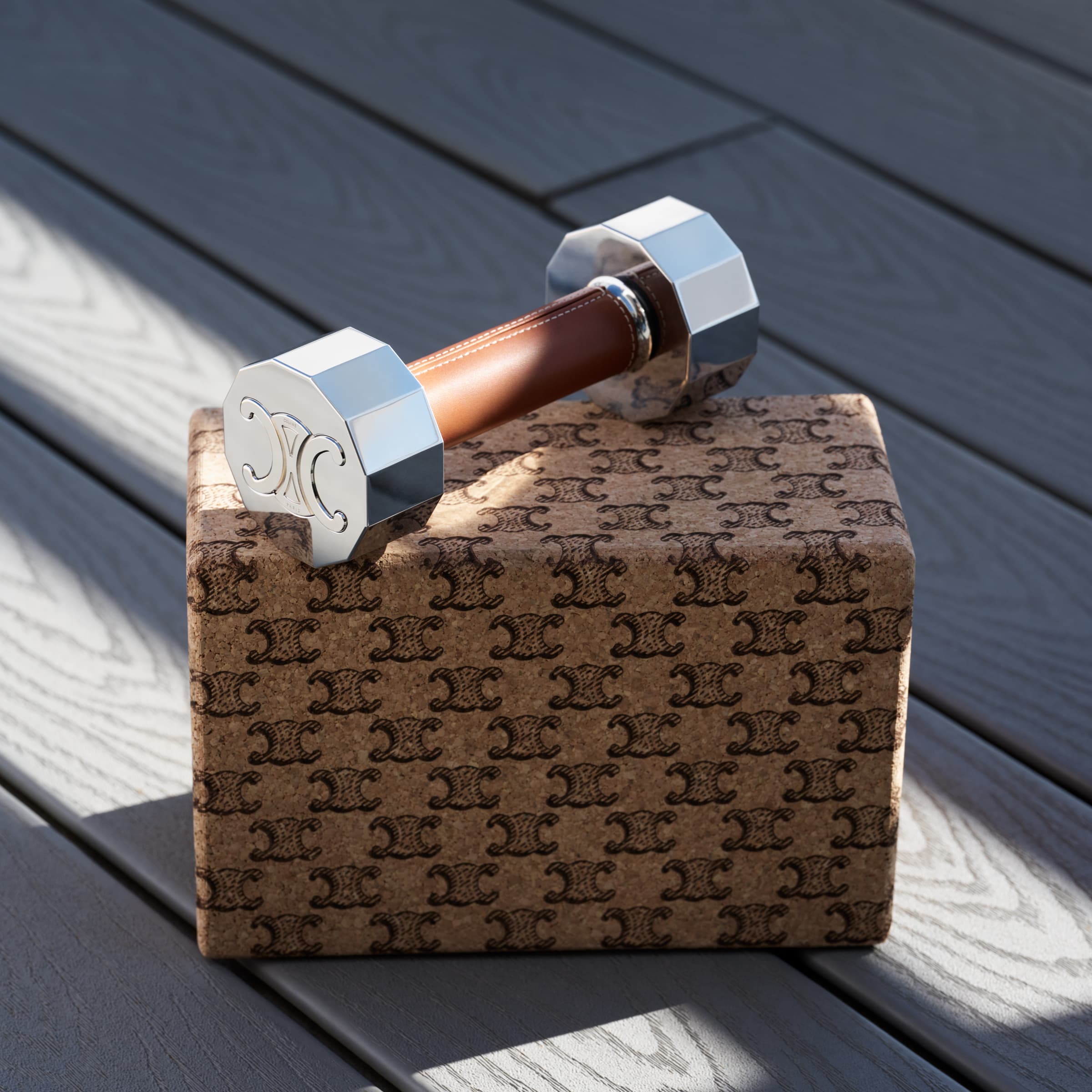 Studio shot of a dumbbell with silver ends and a brown leather handle, resting on a box covered in designer logos on a gray surface.