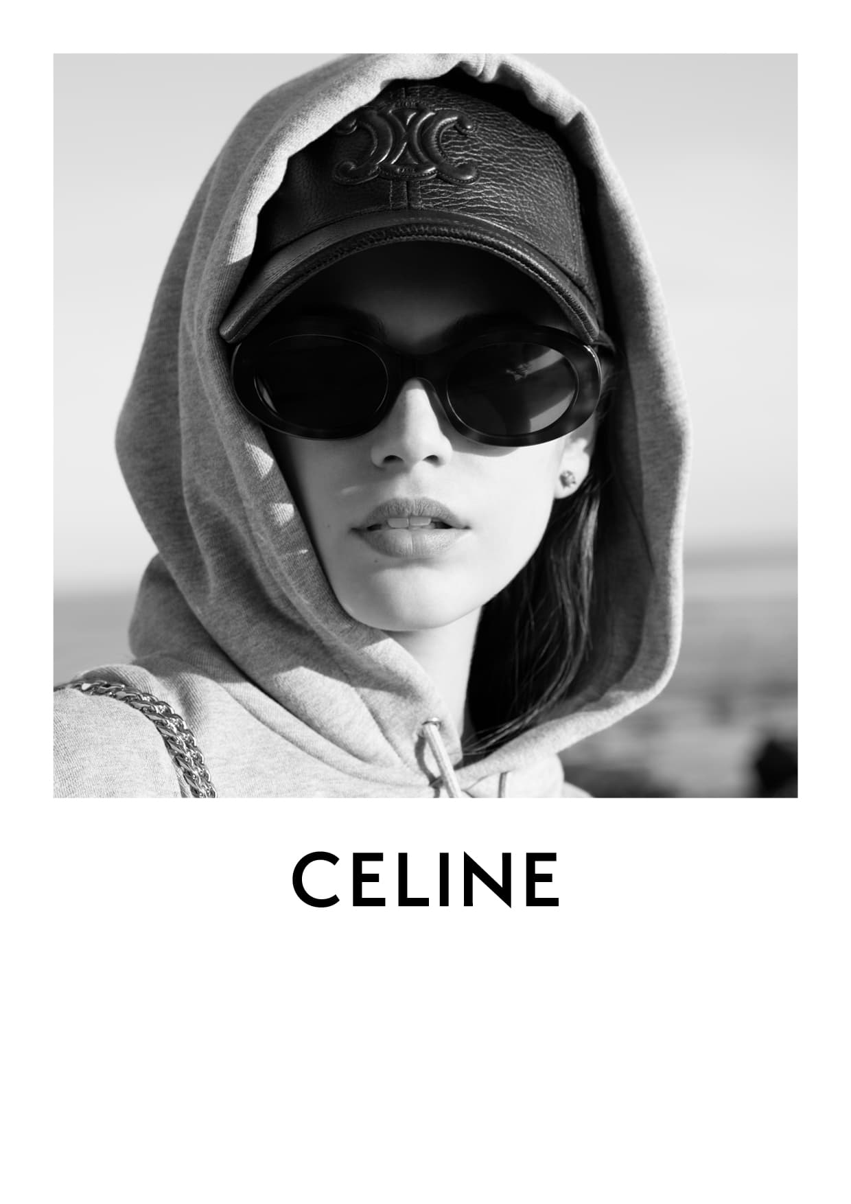 Black-and-white portrait of a woman wearing a baseball cap and hooded sweatshirt, outdoors in daylight, with the Celine logo.