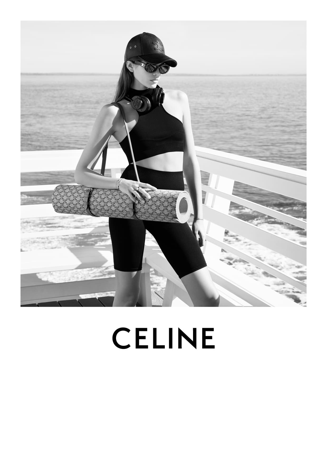 Black and white shot of a woman in workout clothes with a yoga mat and headphones, standing in front of the ocean on a sunny day.