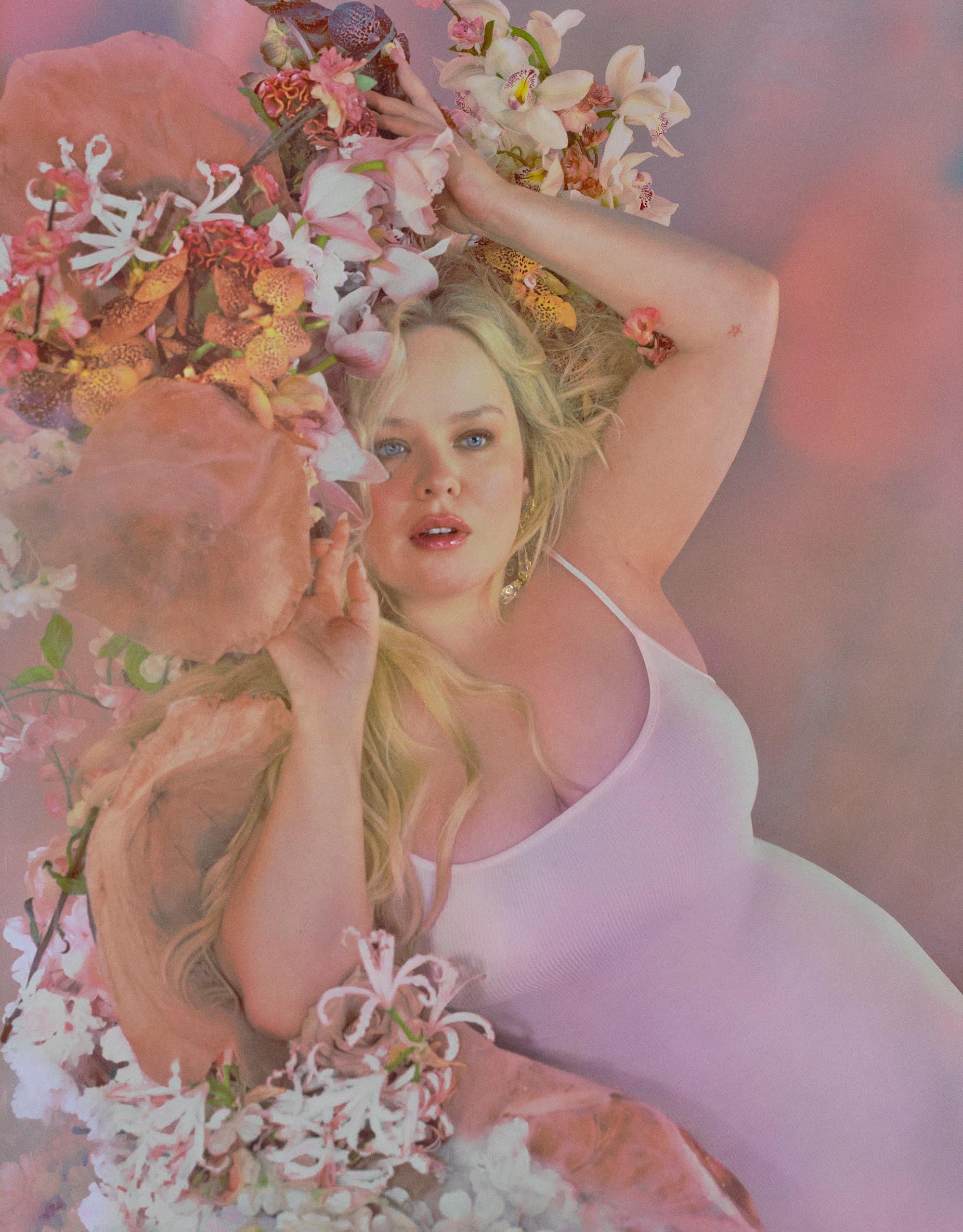 Young woman lies surrounded by pink and white orchids and leaves, wearing a white dress. Soft lighting and pastel tones create a dreamy effect.