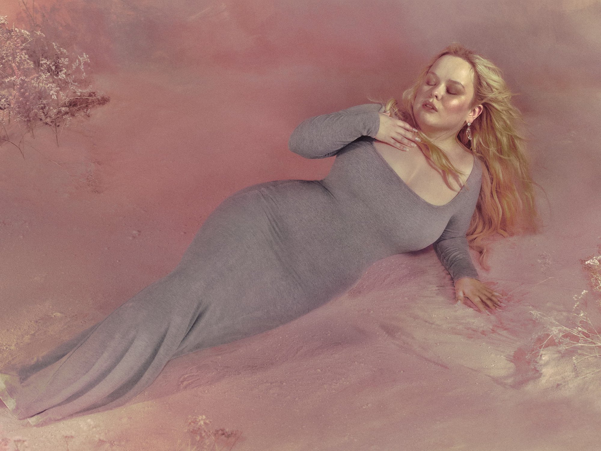 Woman in a long dress lying down in an artistic pose, soft pink background with subtle foliage and artistic lighting.  