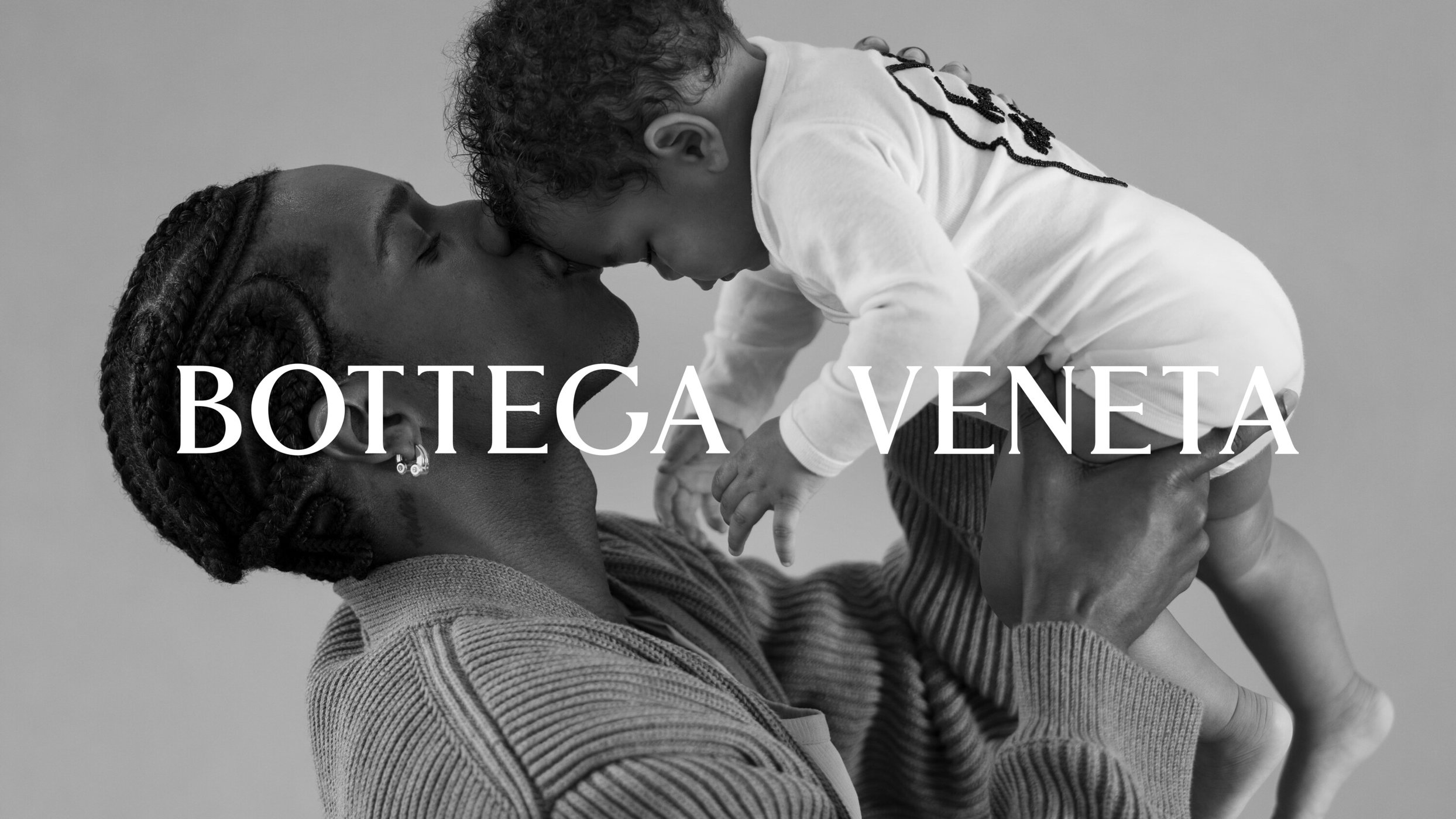 Black and white close-up of a Black man holding a baby, foreheads touching, adorned with Bottega Veneta text overlay, indoor studio shot.