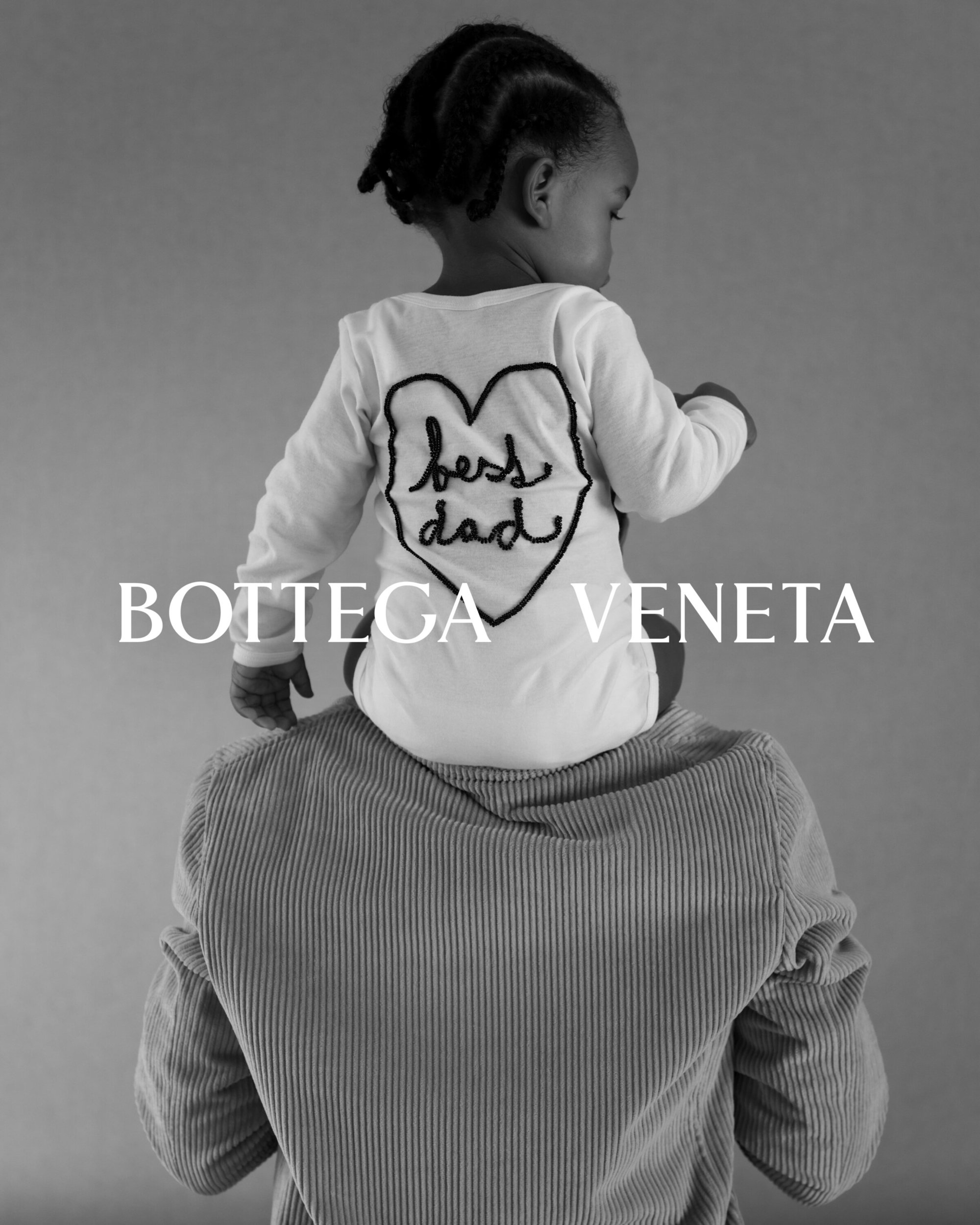 Black-and-white portrait of a toddler riding on an adult's shoulders, both with backs to the camera. The child wears a shirt with 'best dad' on it.