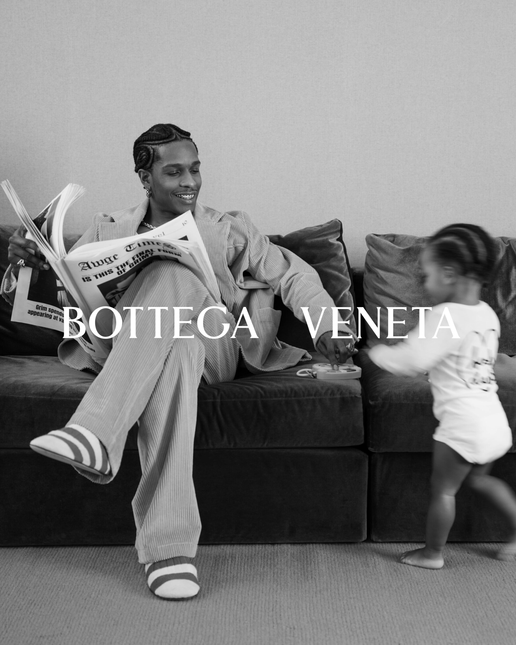 Black and white image of a man reading a newspaper on a couch with a child running near him in an indoor setting.