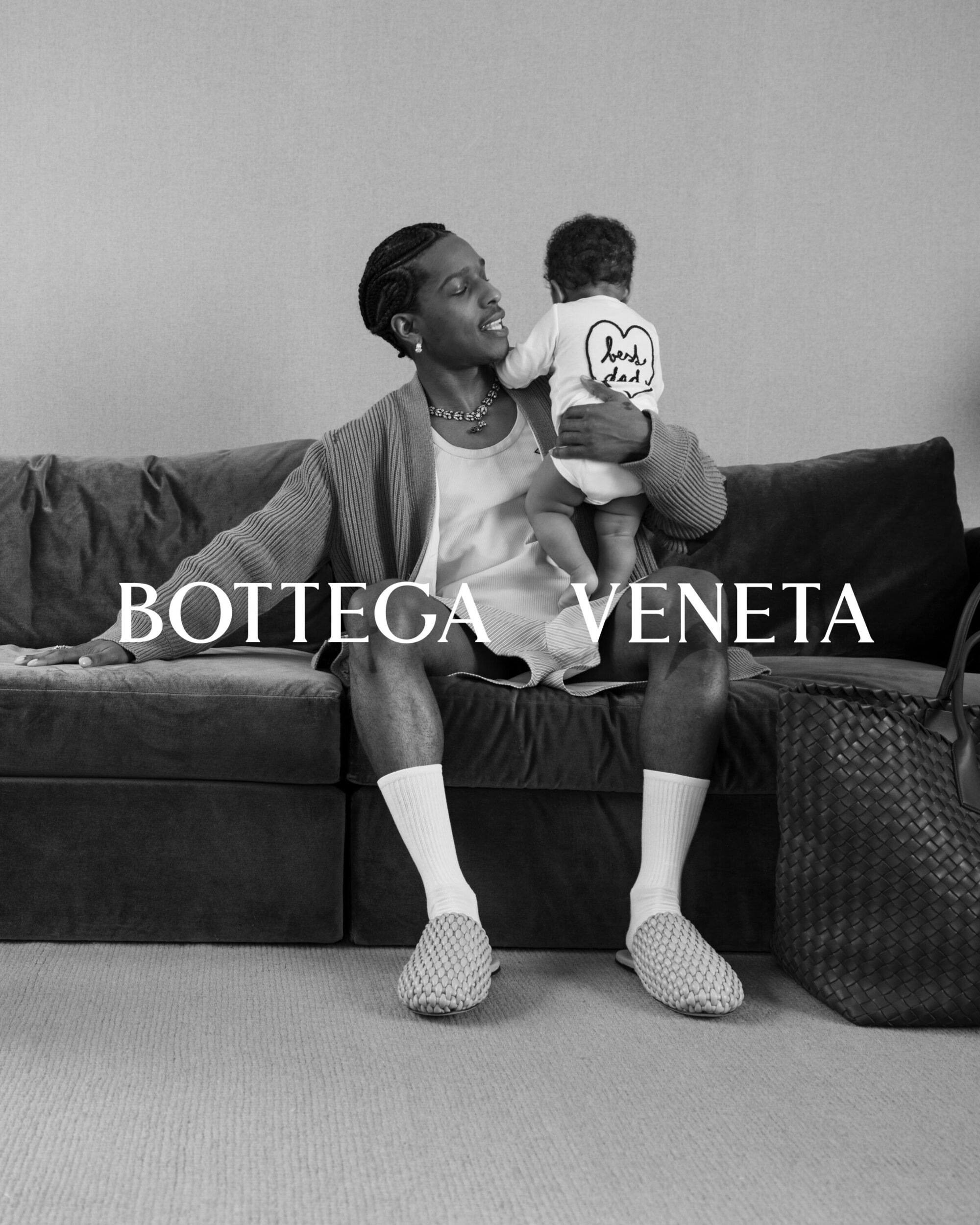 An African American man and a baby are seated on a couch in a black-and-white image, with the words BOTTEGA VENETA superimposed.