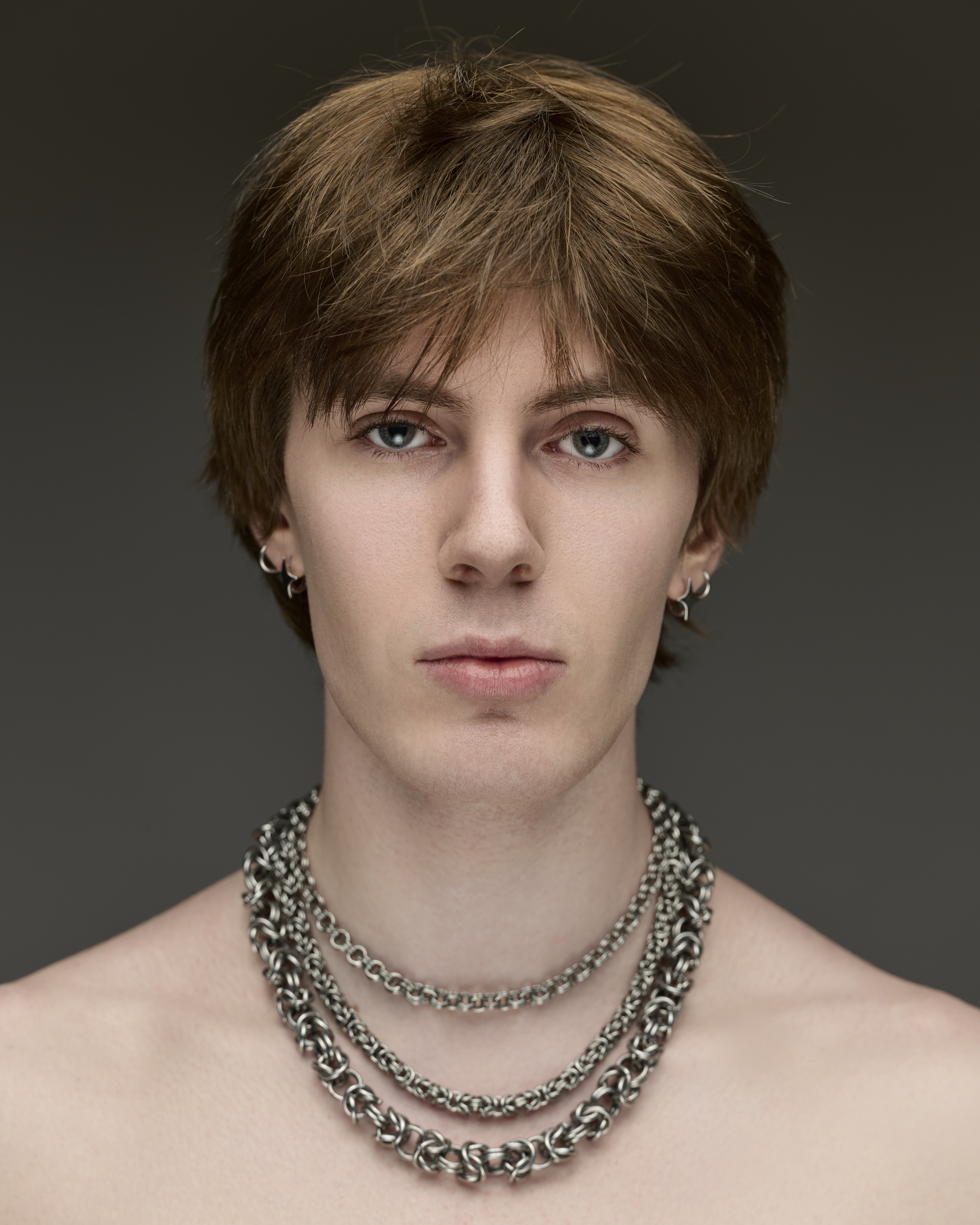 Portrait of a young adult male with auburn hair and blue eyes, wearing three layered silver necklaces against a grey background.