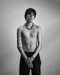 Monochrome studio portrait of a young man wearing a chain necklace and dark textured trousers against a neutral background.