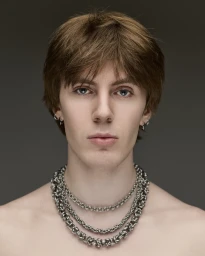 Close-up portrait of a young man with brown hair wearing layered silver chain necklaces against a grey background.