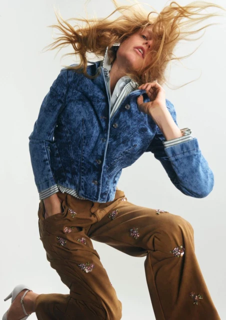 Woman in denim jacket and embellished pants jumps in a studio. Fashion shot with blonde hair movement, a light background, and neutral color palette.