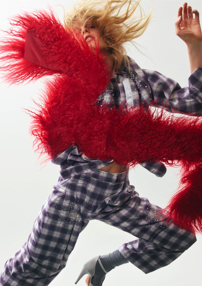 Fashionable blonde woman in mid-air, sporting a plaid outfit and heels, accessorized with a striking red faux fur scarf in a studio setting.