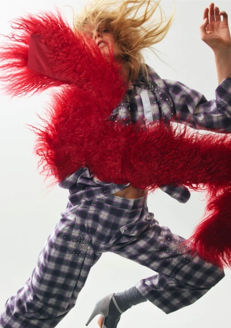 Fashionable blonde woman in mid-air, sporting a plaid outfit and heels, accessorized with a striking red faux fur scarf in a studio setting.