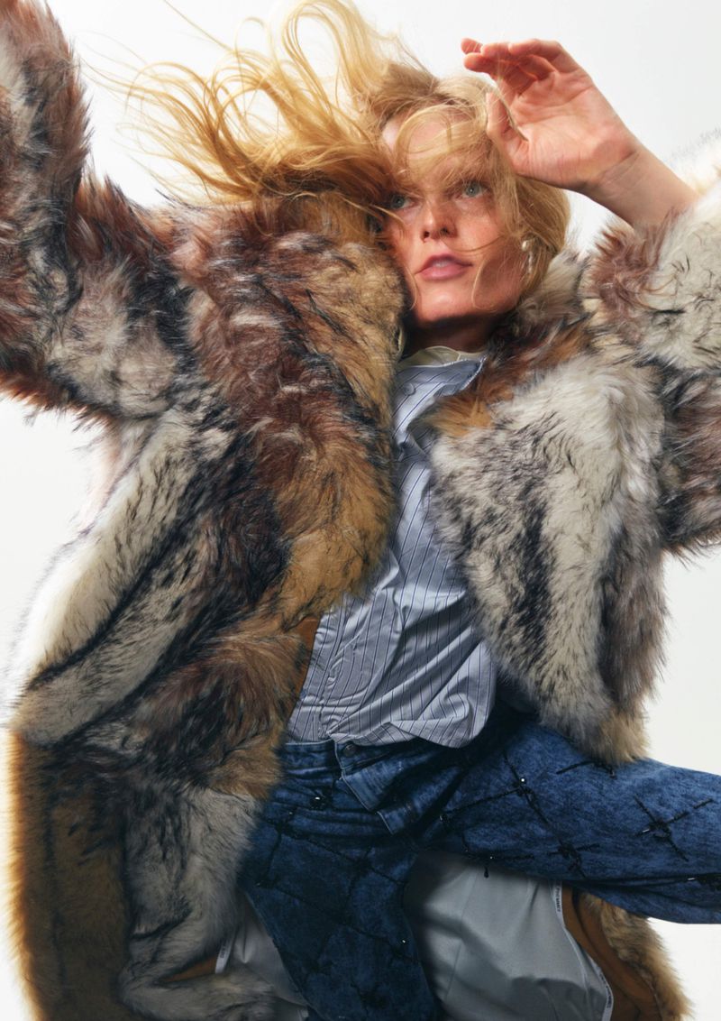 Fashionable woman in motion, wearing a fur coat, blue jeans, and light shirt against a bright background. Wind blown hair adds to the dynamic feel.