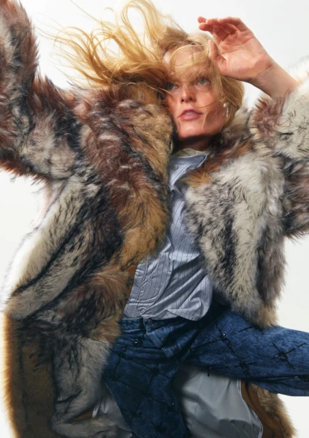 Fashionable woman in motion, wearing a fur coat, blue jeans, and light shirt against a bright background. Wind blown hair adds to the dynamic feel.