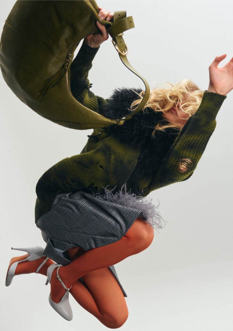 A woman jumps in a green, black, and gray outfit with a green purse and gray heels against a white background in a fashion shot.