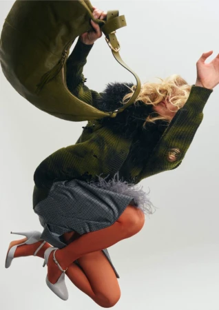 A woman jumps in a green, black, and gray outfit with a green purse and gray heels against a white background in a fashion shot.