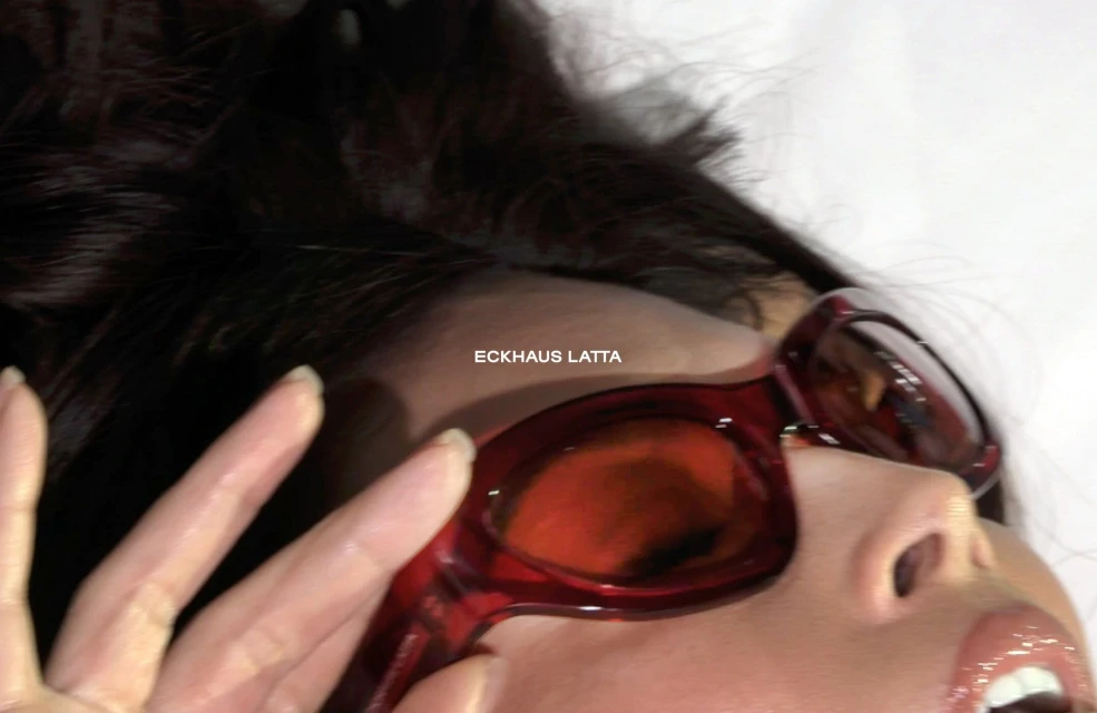 Brunette woman wearing red-framed sunglasses in an extreme close-up, face partially visible with a light complexion and glossy lips.