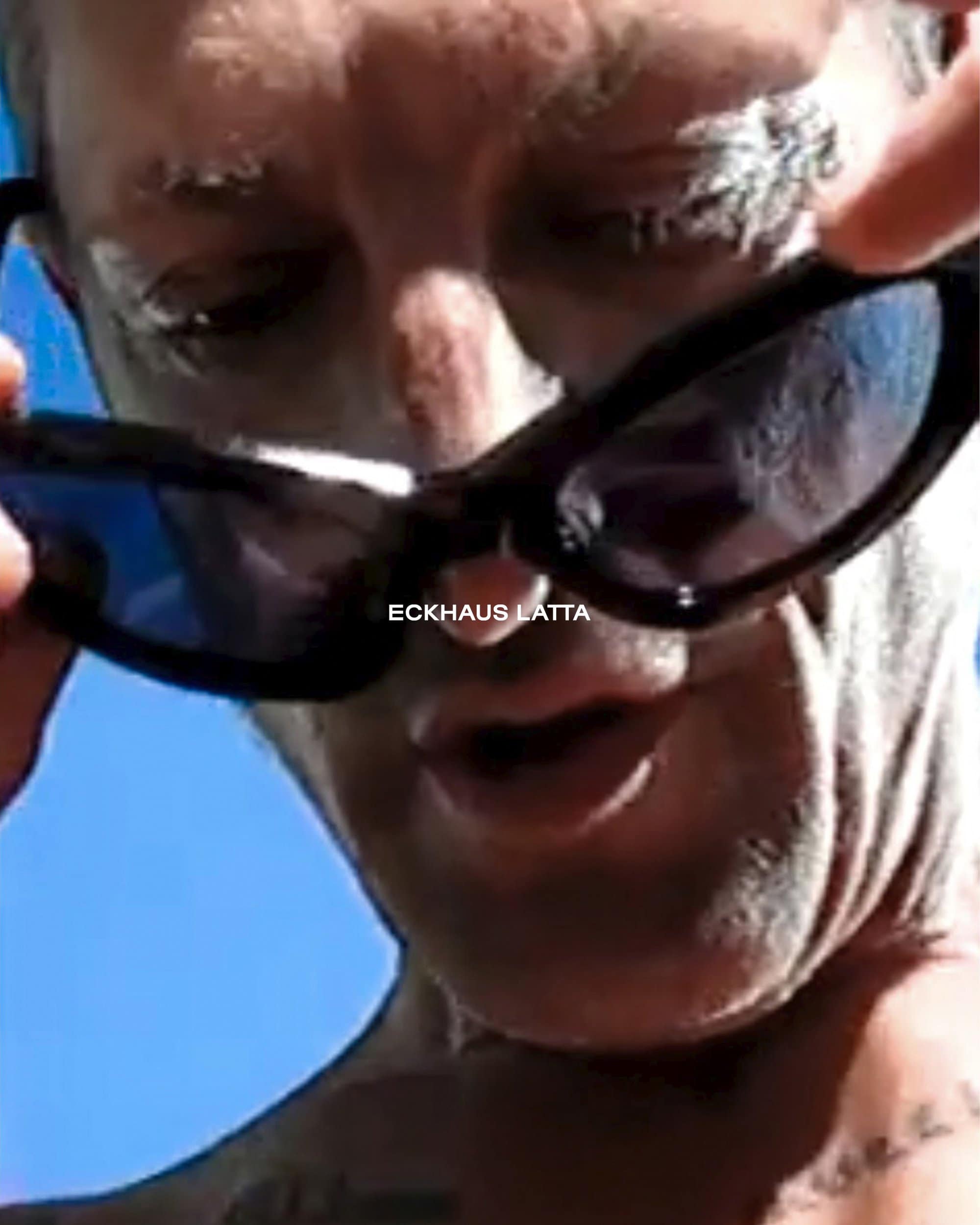 Adult male with sunglasses at eye level, outside with blue sky background. Clear facial features with tattoos visible on skin.