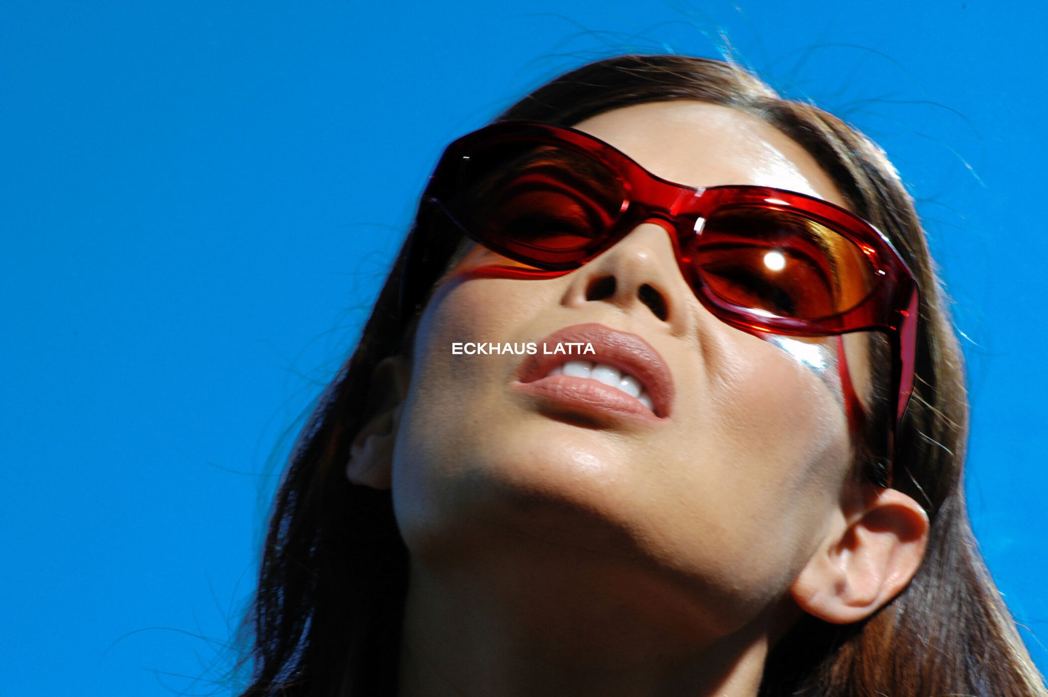 Portrait of a woman with red sunglasses looking up against a bright blue sky, creating a striking contrast and modern aesthetic.