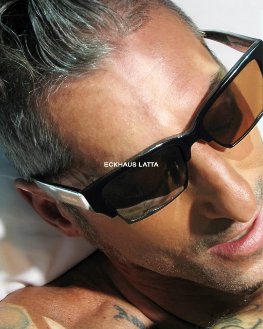 Close-up of a reflective sunglass lens on a male model, fair skin, with silver hair and a tattoo on the neck and chest.