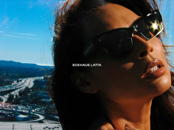 Close-up of a woman wearing sunglasses outdoors against a cityscape backdrop and bright blue sky, fashion brand Eckhaus Latta displayed.