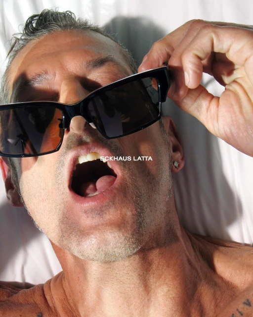Close-up portrait of a man with an open mouth holding sunglasses up to his face against a white background with branding in the foreground.
