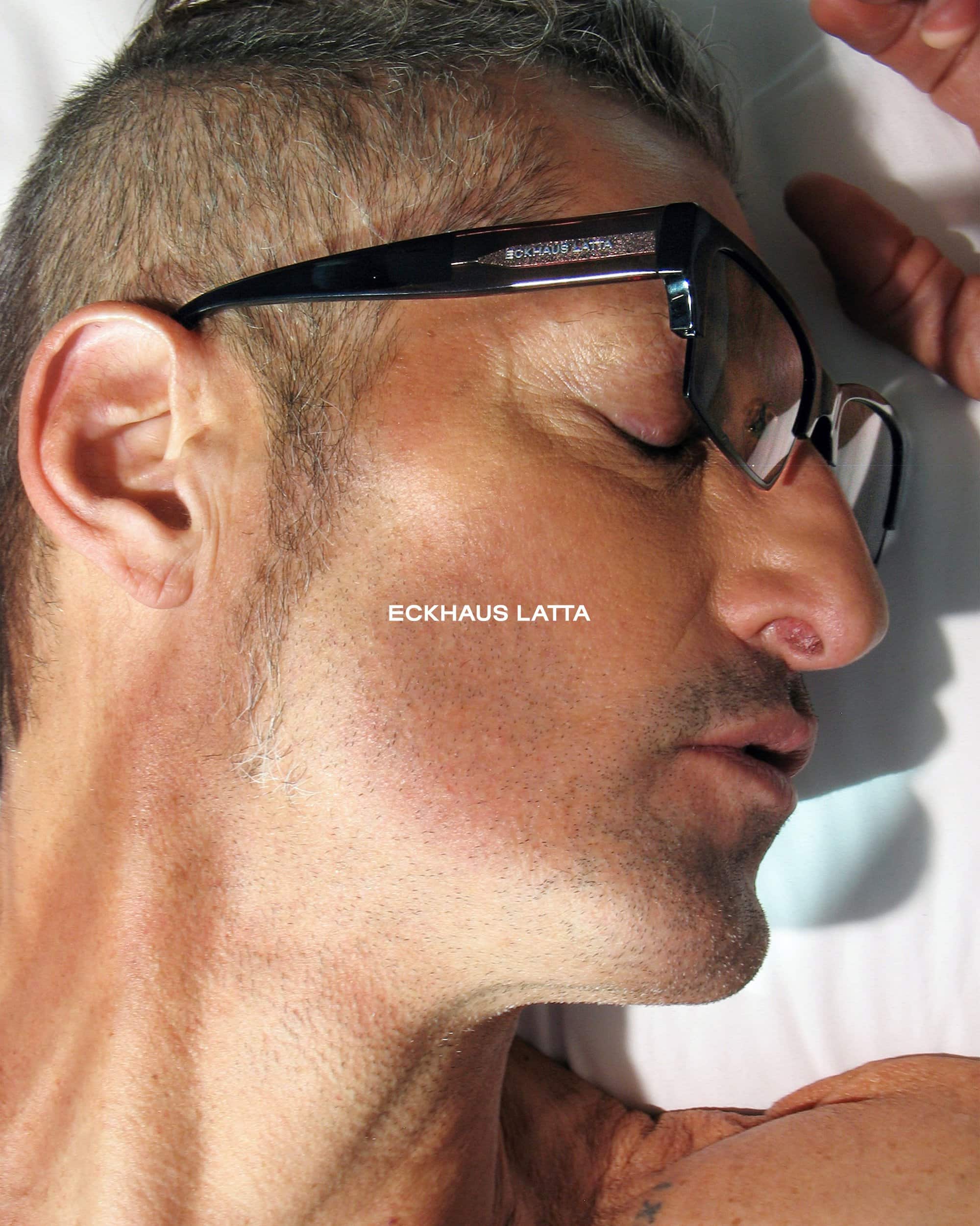 Profile of a light-skinned adult male with short grey hair, wearing black eyeglasses, laying on a white surface in soft lighting.
