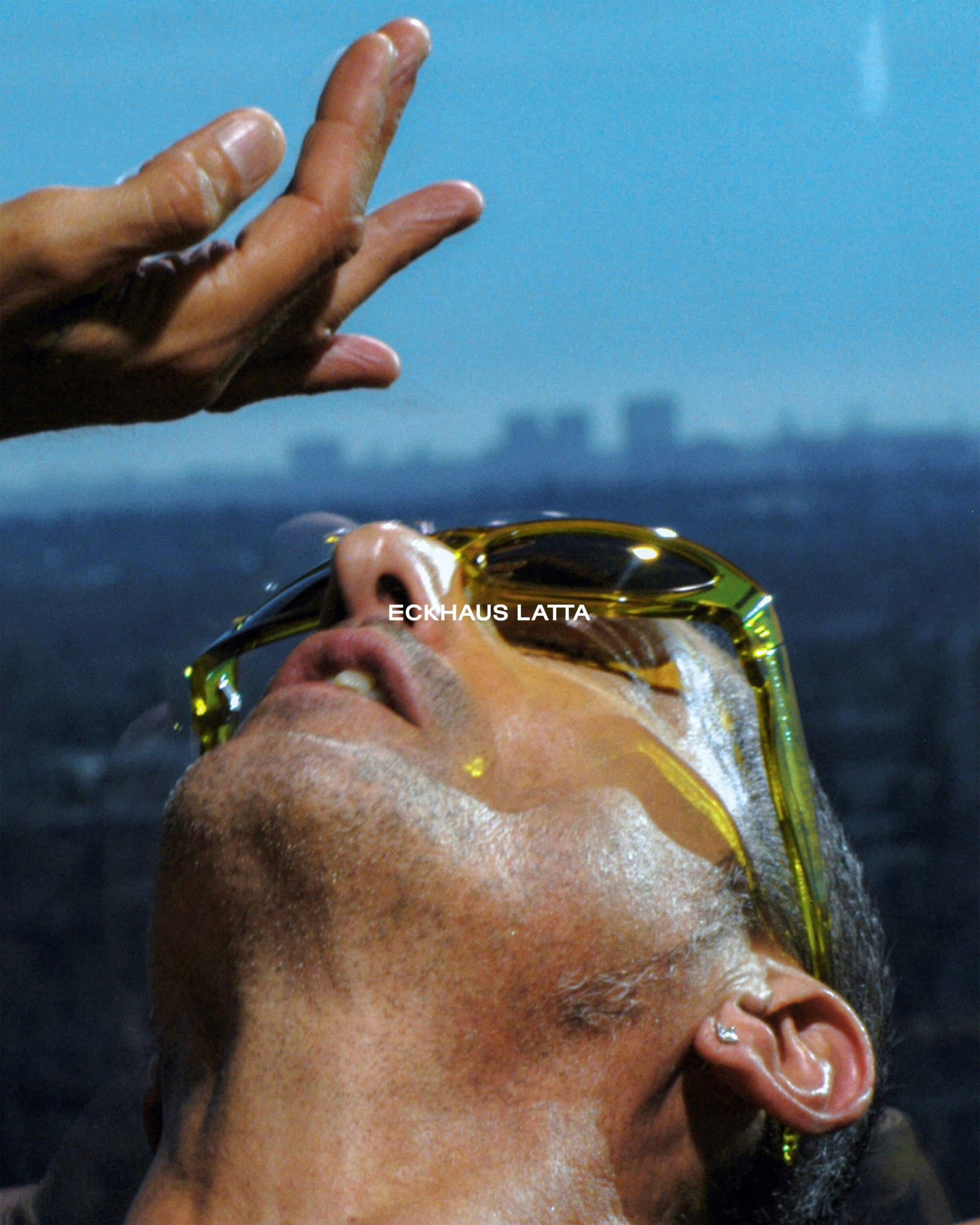 Portrait of adult male looking up and wearing sunglasses with liquid dripping down, set against a city skyline and blue sky.
