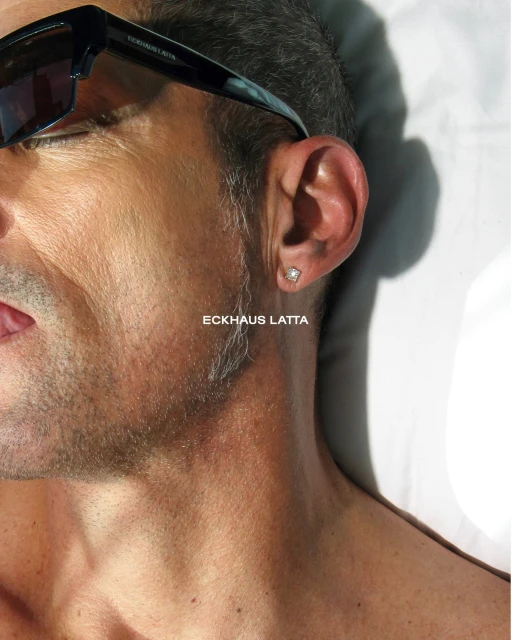 Close-up of an adult male with sunglasses, highlighting his ear with an earring, fair skin, and the brand name Eckhaus Latta visible.