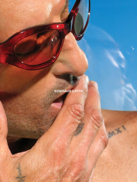 Close-up of a male face wearing red-framed sunglasses, with his hand near his mouth, set against a bright blue sky background.