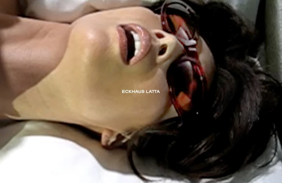 Close-up of a woman with dark hair, wearing sunglasses, lying down, with her mouth open in an indoor setting. Fashion and beauty.
