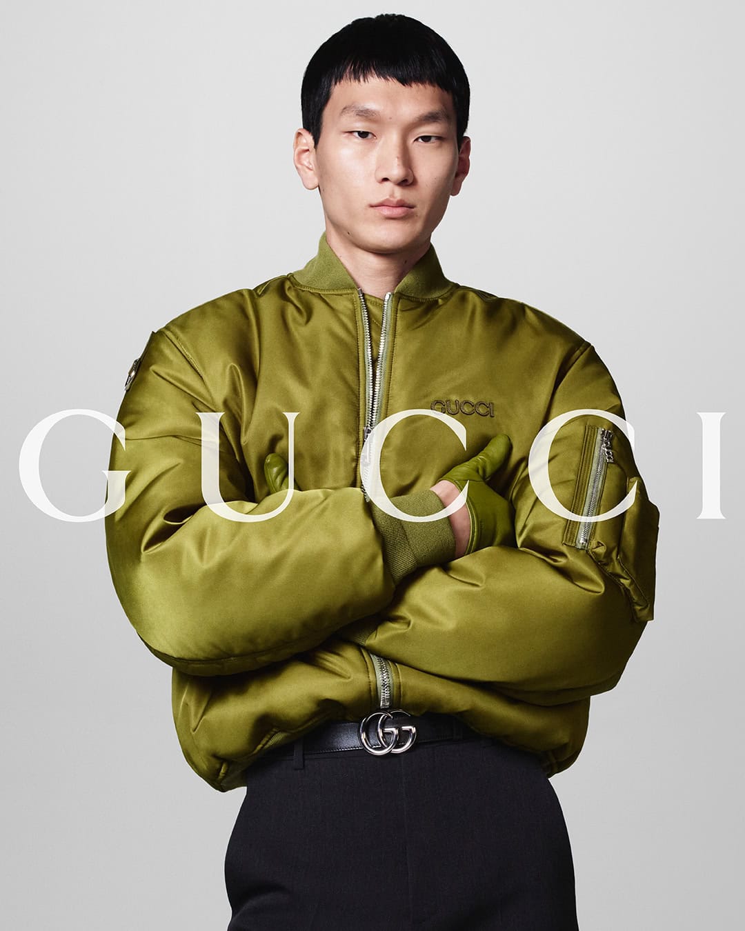 Asian male model poses in a studio wearing a green Gucci bomber jacket with black pants and a belt. Gucci logo is in the background.