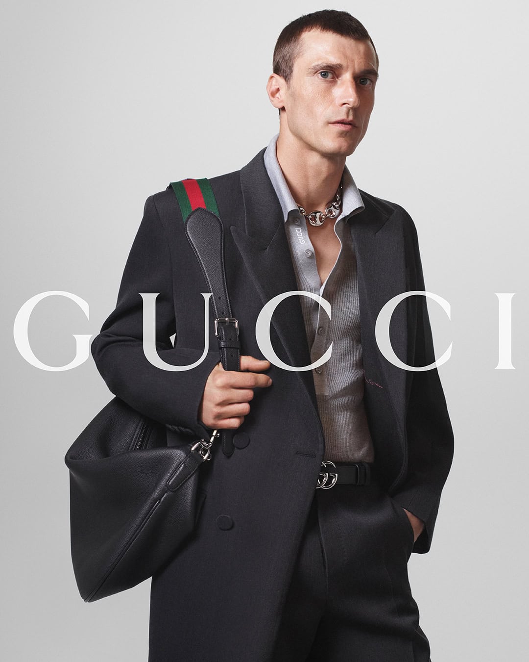 A man in a dark suit holds a black Gucci bag with a green and red strap. The Gucci logo is visible in the background.