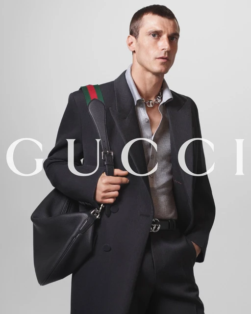 A man in a dark suit holds a black Gucci bag with a green and red strap. The Gucci logo is visible in the background.