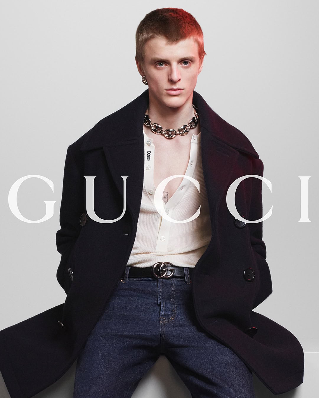 A fair-skinned male model poses wearing a black overcoat, cardigan, jeans, jewelry, and belt in a studio setting for a fashion brand ad.