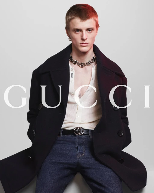 A fair-skinned male model poses wearing a black overcoat, cardigan, jeans, jewelry, and belt in a studio setting for a fashion brand ad.