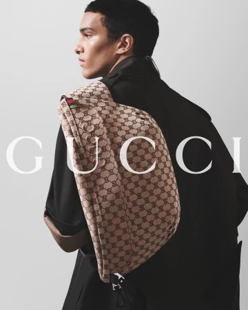 Male fashion model poses with a Gucci messenger bag slung over his shoulder in a studio environment with neutral, soft lighting.