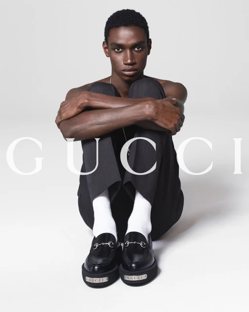 Fashion shot of an adult male sitting with legs bent, wearing white socks and black Gucci loafers against a white background.